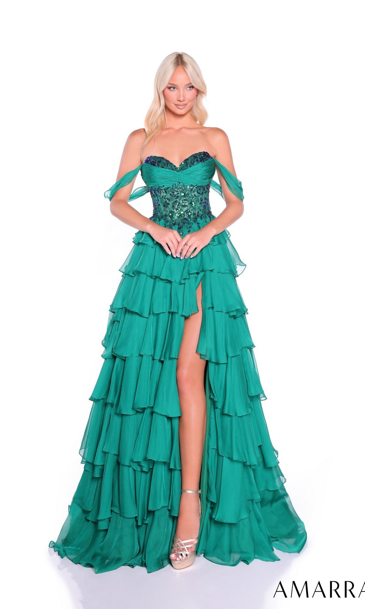 A woman wears the Amarra 89313 long prom dress—an off-shoulder emerald green gown with a chiffon tiered skirt, beaded bodice, thigh-high slit—and silver high heels, posing against a white background.